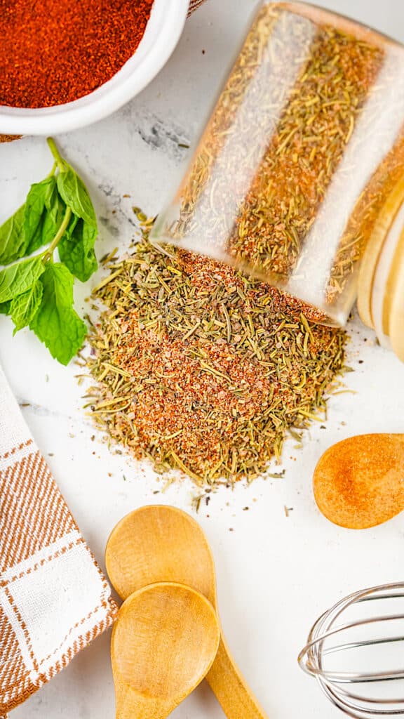 Turkey Rub spilling out of a clear jar onto a white marble table next to a wooden spoons and a whisk