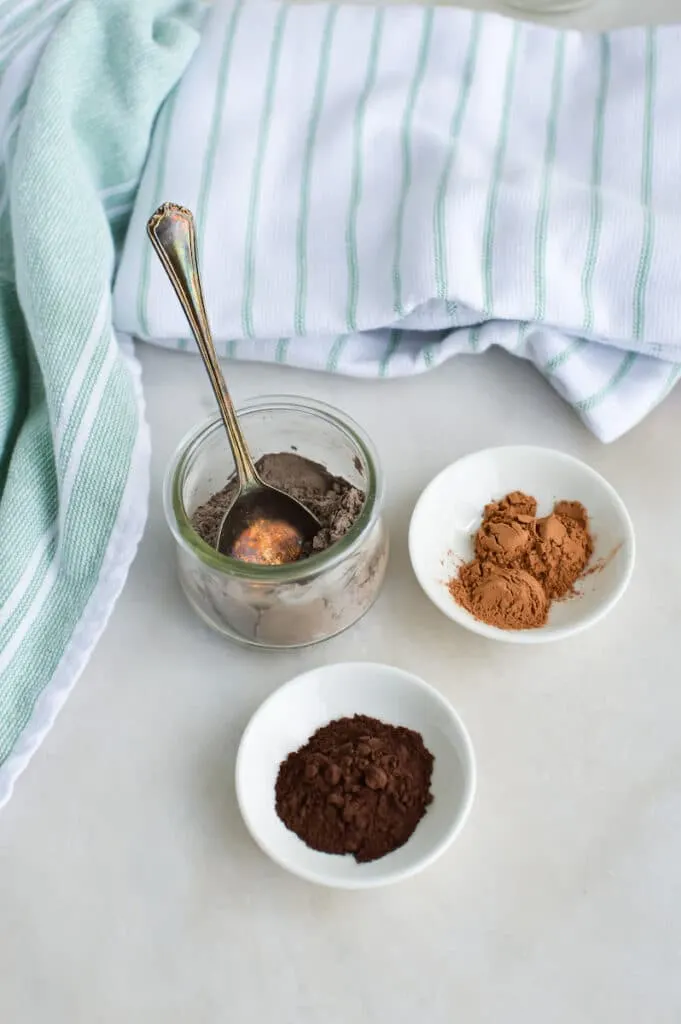 Two bowls of cocoa powder and a spoon on a white table for DIY Dry Shampoo for dark hair.