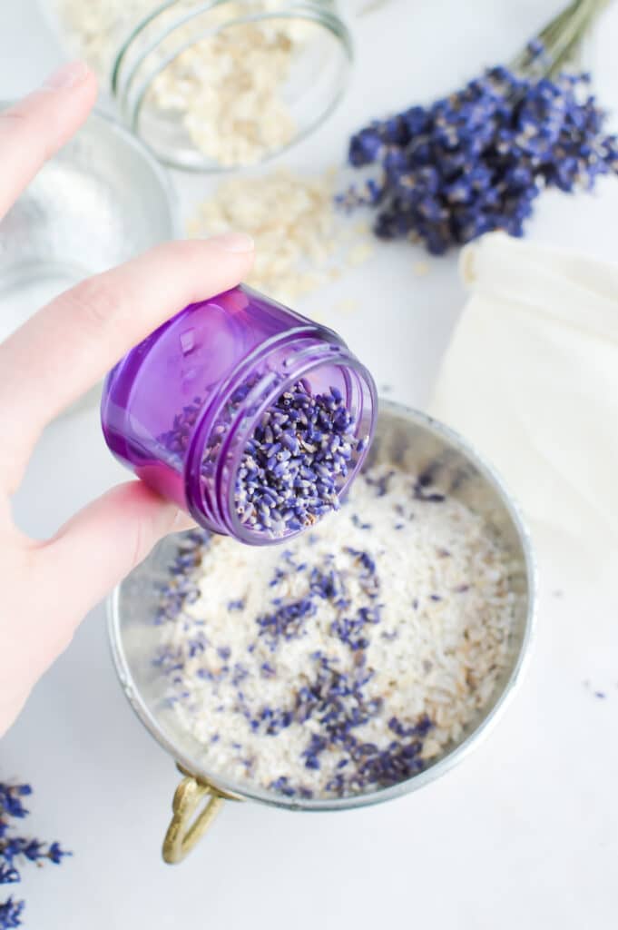 A hand is sprinkling lavender into a bowl of oats making Oat Bath Soak.